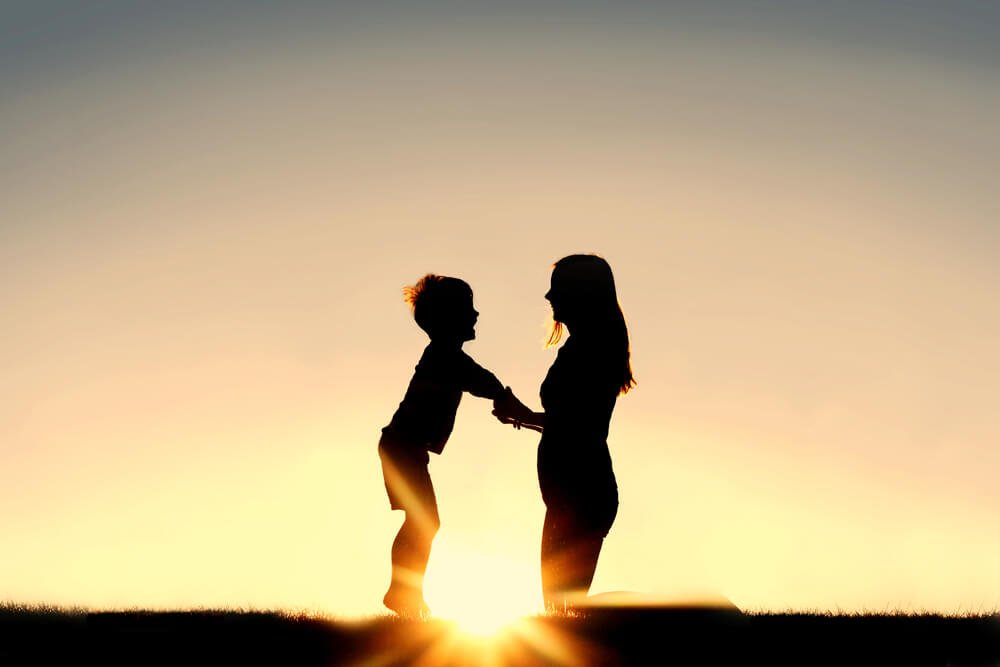 Silhouette of a young mother lovingly holding hands with her happy little child outside in front of a sunset in the sky.