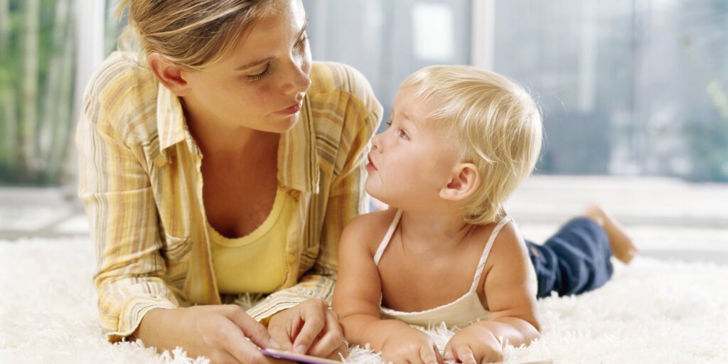 Woman and female toddler (21-24 months) lying on rug with book, looking at each other