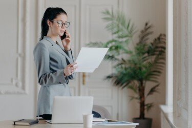 Serious brunette female employee stands near desktop, holds paper documents, talks via smartphone, wears formal clothes and spectacles, discusses business strategy with partner, organizes meeting